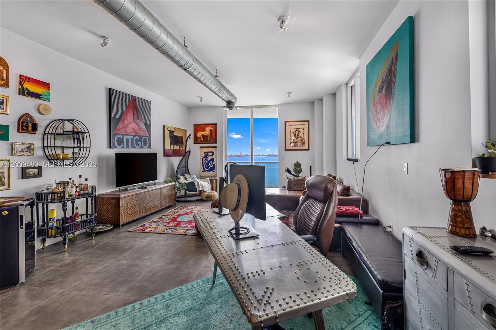 A modern living room features a metal desk, industrial ceiling ductwork, and a waterfront view — 700 NE 25th St # 1104, Miami FL 33137, MIAMI, FL