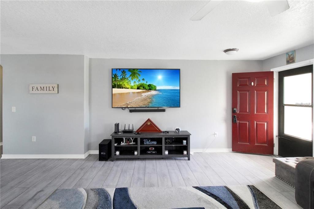 A living room features a wall-mounted television, a media console, and a red front door — 15427 WINTER BREEZE LANE, WIMAUMA, FL