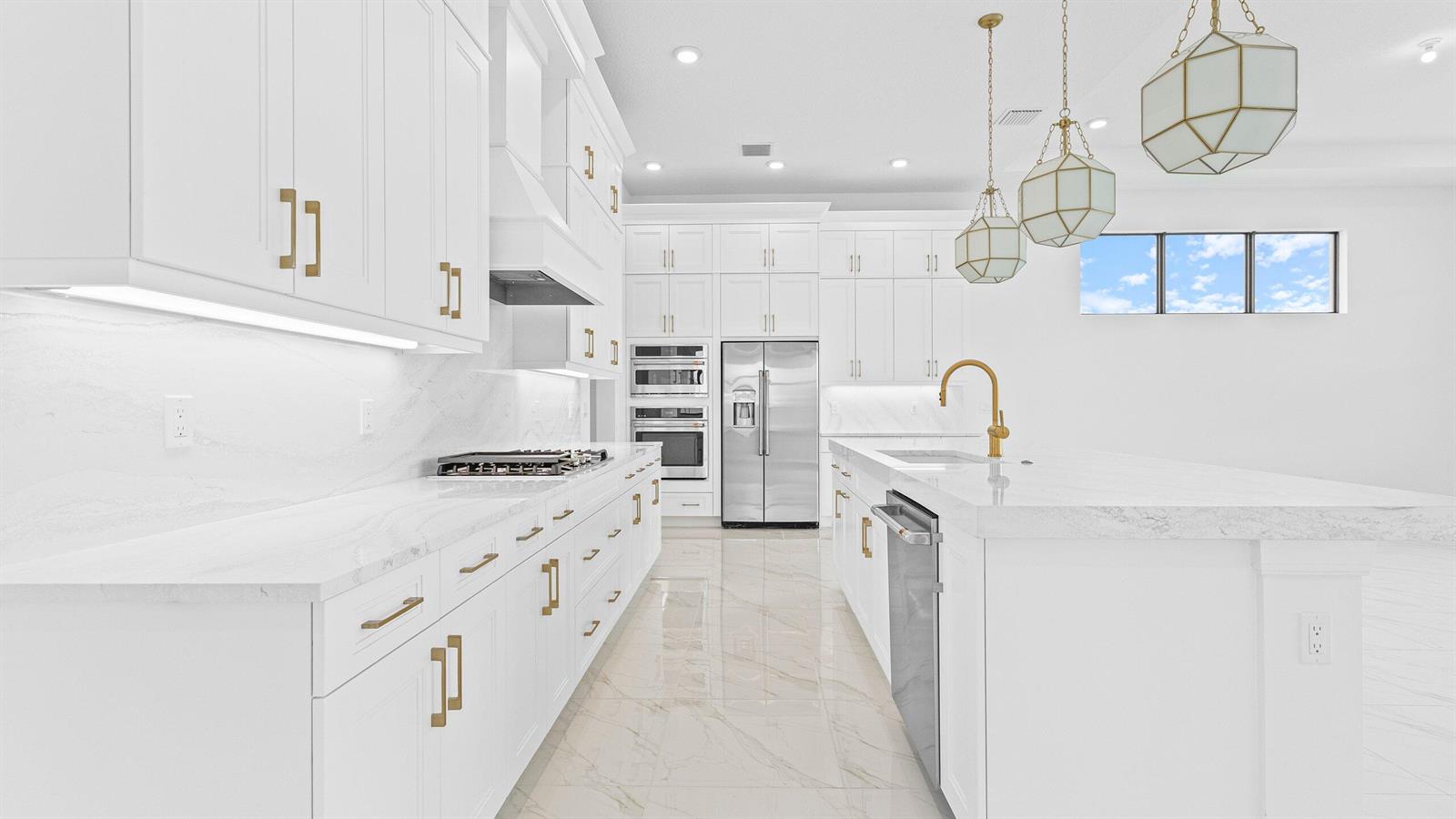 A modern white kitchen features marble countertops, gold hardware, stainless steel appliances, and pendant lighting — 5474 Hutchinson Way, FL 33470, FL