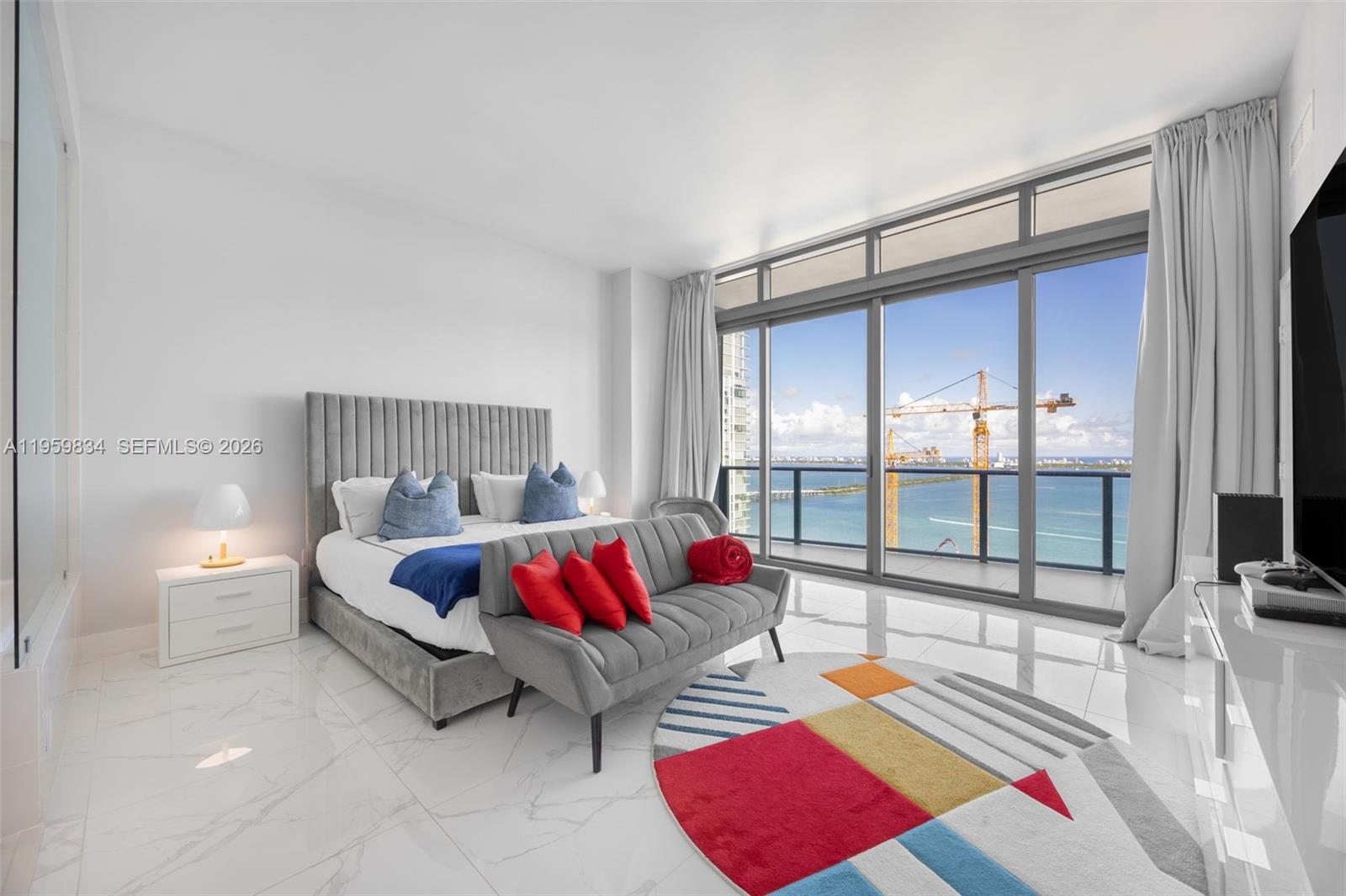 A modern bedroom features a gray tufted bed, a sofa, and floor-to-ceiling windows overlooking water — 460 NE 28th St # 4002, Miami FL 33137, MIAMI, FL