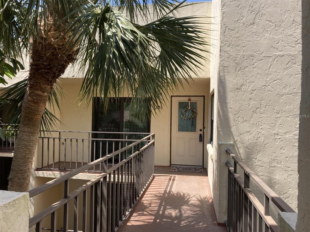 An exterior view of an apartment entrance with a walkway, metal railings, and a palm tree — 6430 SUN EAGLE LANE UNIT 203, BRADENTON, FL