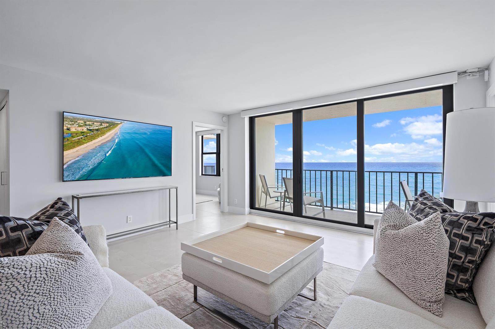 A living room features a wall-mounted television, a central ottoman, and a balcony overlooking ocean — 3456 S Ocean Boulevard # 701, Palm Beach FL 33480, PALM BEACH, FL