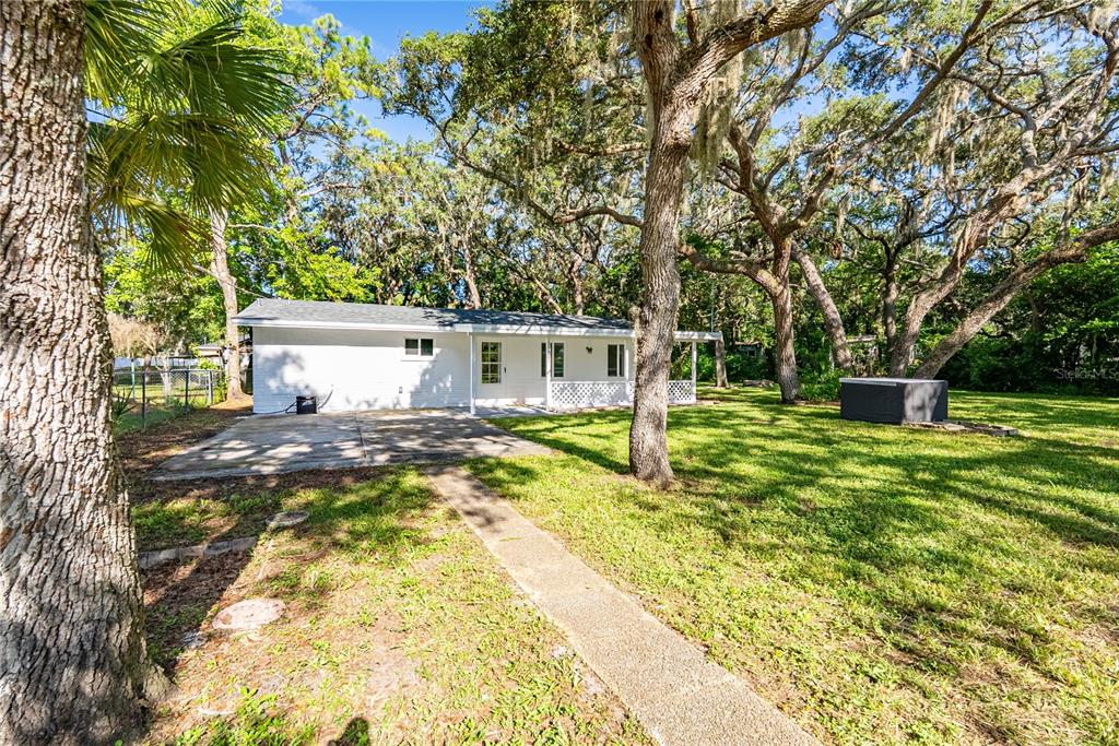 The exterior of a white house features a concrete patio, a walkway, and large oak trees — 11303 CHICAGO AVENUE, NEW PORT RICHEY, FL