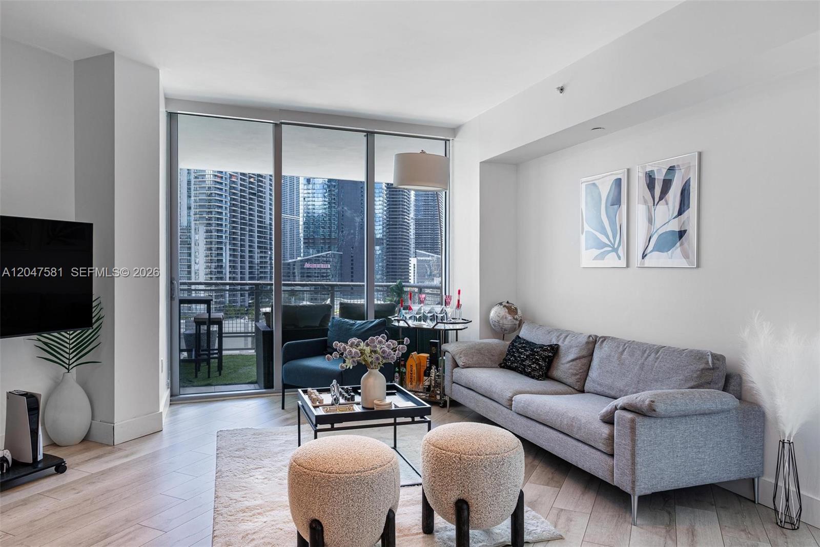 A modern living room features a grey sofa, two ottomans, and floor-to-ceiling windows overlooking skyscrapers — 350 S Miami Ave # 2108, Miami FL 33130, MIAMI, FL