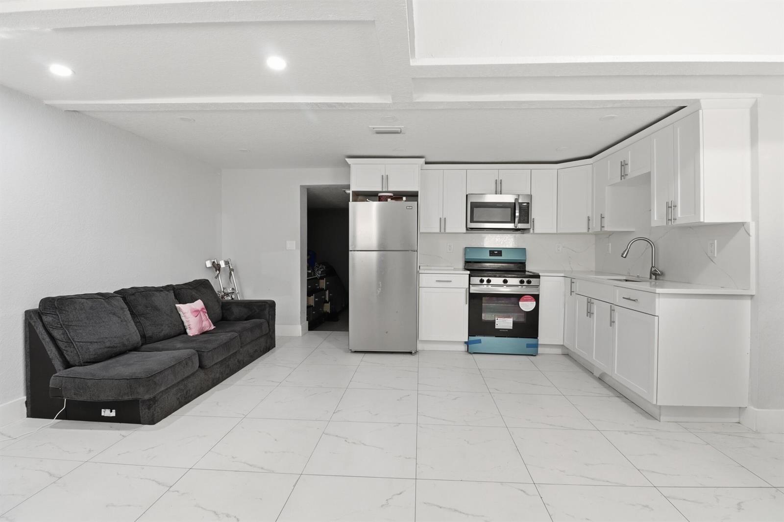 A modern kitchen with white cabinets and stainless steel appliances sits beside a dark sofa — 1152 Sunset Road, West Palm Beach FL 33406, WEST PALM BEACH, FL