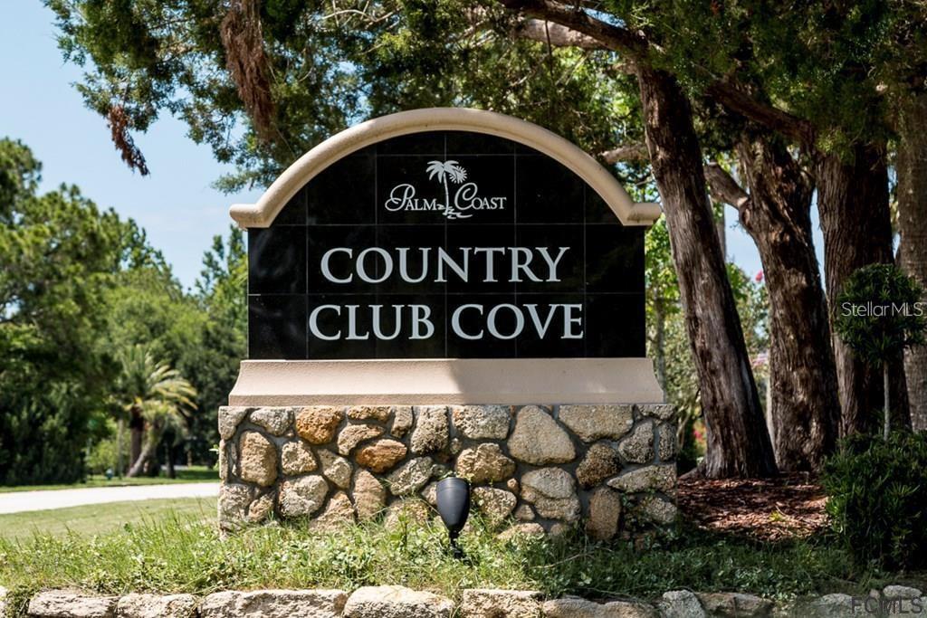 A stone-based monument sign for Country Club Cove stands outdoors among trees — 48 CLUB HOUSE DRIVE UNIT 106, PALM COAST, FL