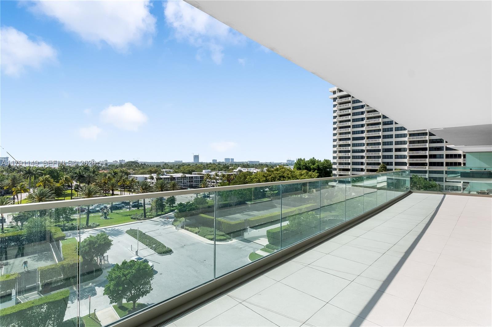 A spacious balcony with glass railings overlooks a parking area, tennis courts, and distant buildings — 10201 Collins Ave # 605, Bal Harbour FL 33154, BAL HARBOUR, FL
