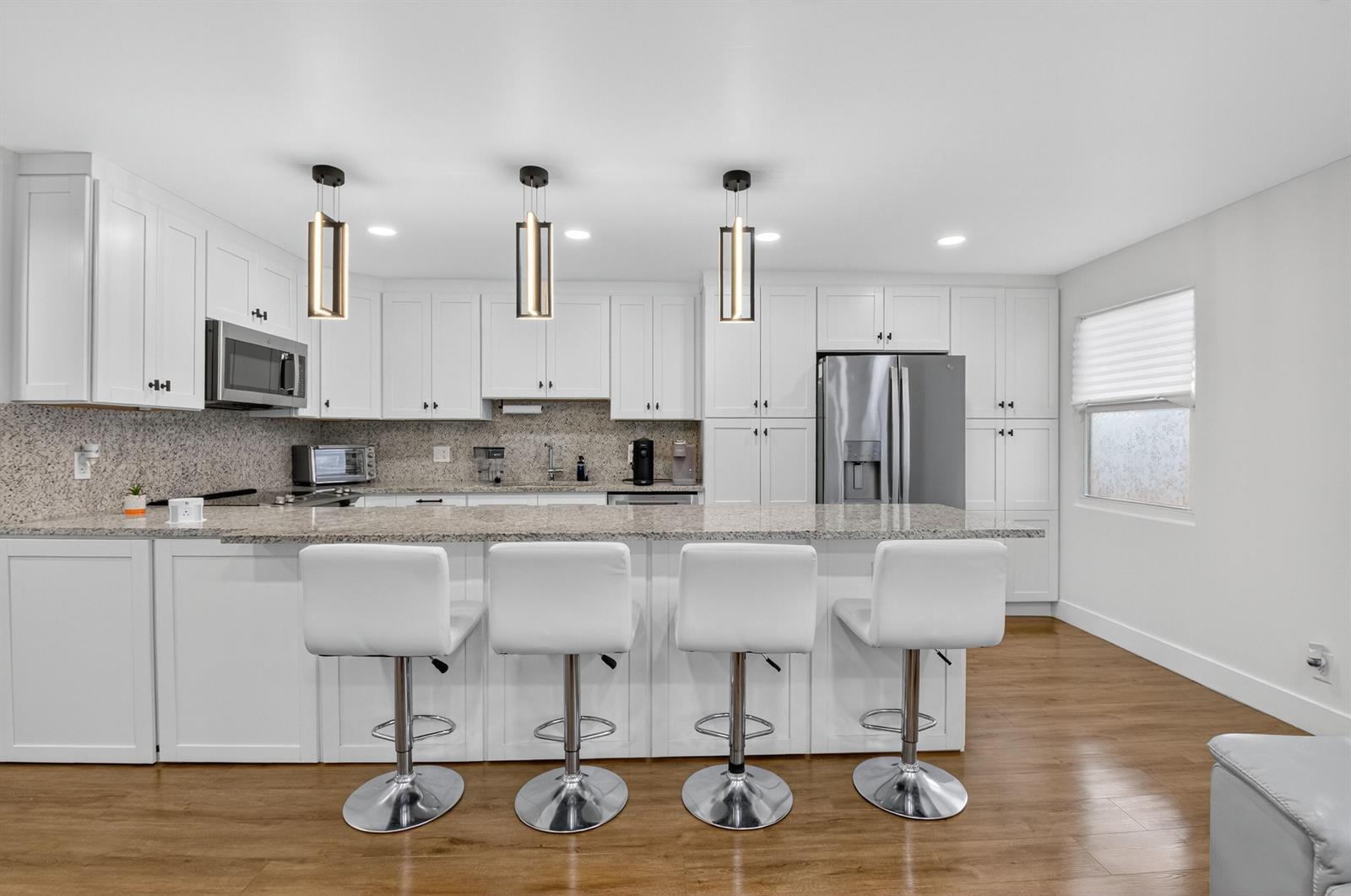 A modern kitchen features white cabinetry, granite countertops, a breakfast bar, and four white stools — 3005 Guildford A, Boca Raton FL 33434, BOCA RATON, FL