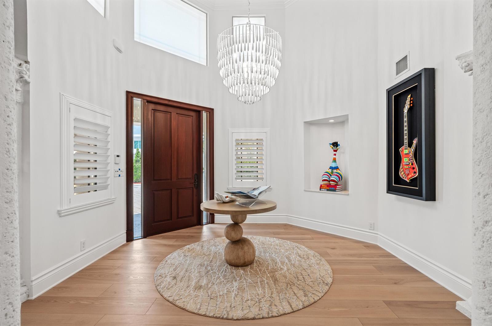 A bright foyer features a wooden front door, a chandelier, and a framed guitar display — 1098 Spanish River Road, Boca Raton FL 33432, BOCA RATON, FL