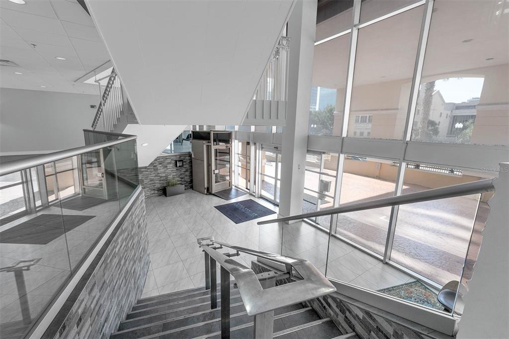 A modern lobby features a glass-railed staircase, tiled floors, and floor-to-ceiling windows overlooking a plaza — 151 E WASHINGTON STREET UNIT 404, ORLANDO, FL