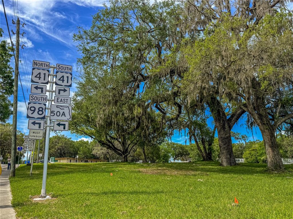 A grassy lot features large oak trees and a metal signpost with multiple highway markers — 615 OLD HOSPITAL DRIVE, BROOKSVILLE, FL