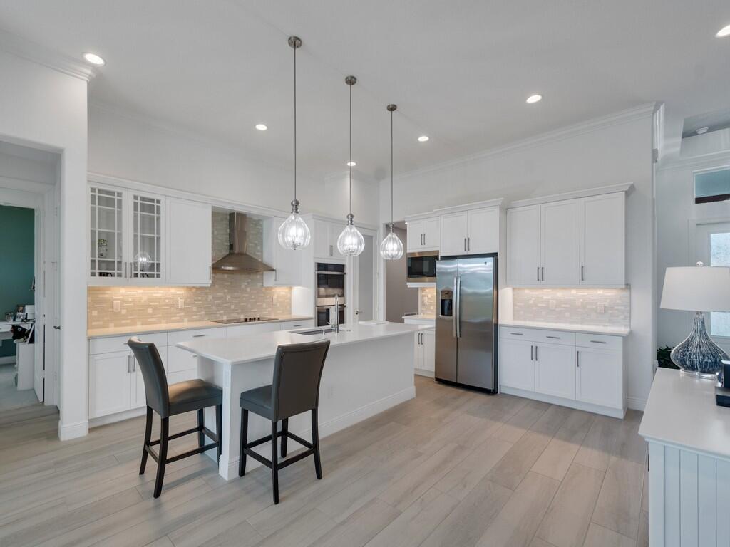 A modern white kitchen features a central island, stainless steel appliances, and light wood flooring — 8220 Halbert Lane, Vero Beach FL 32968, VERO BEACH, FL