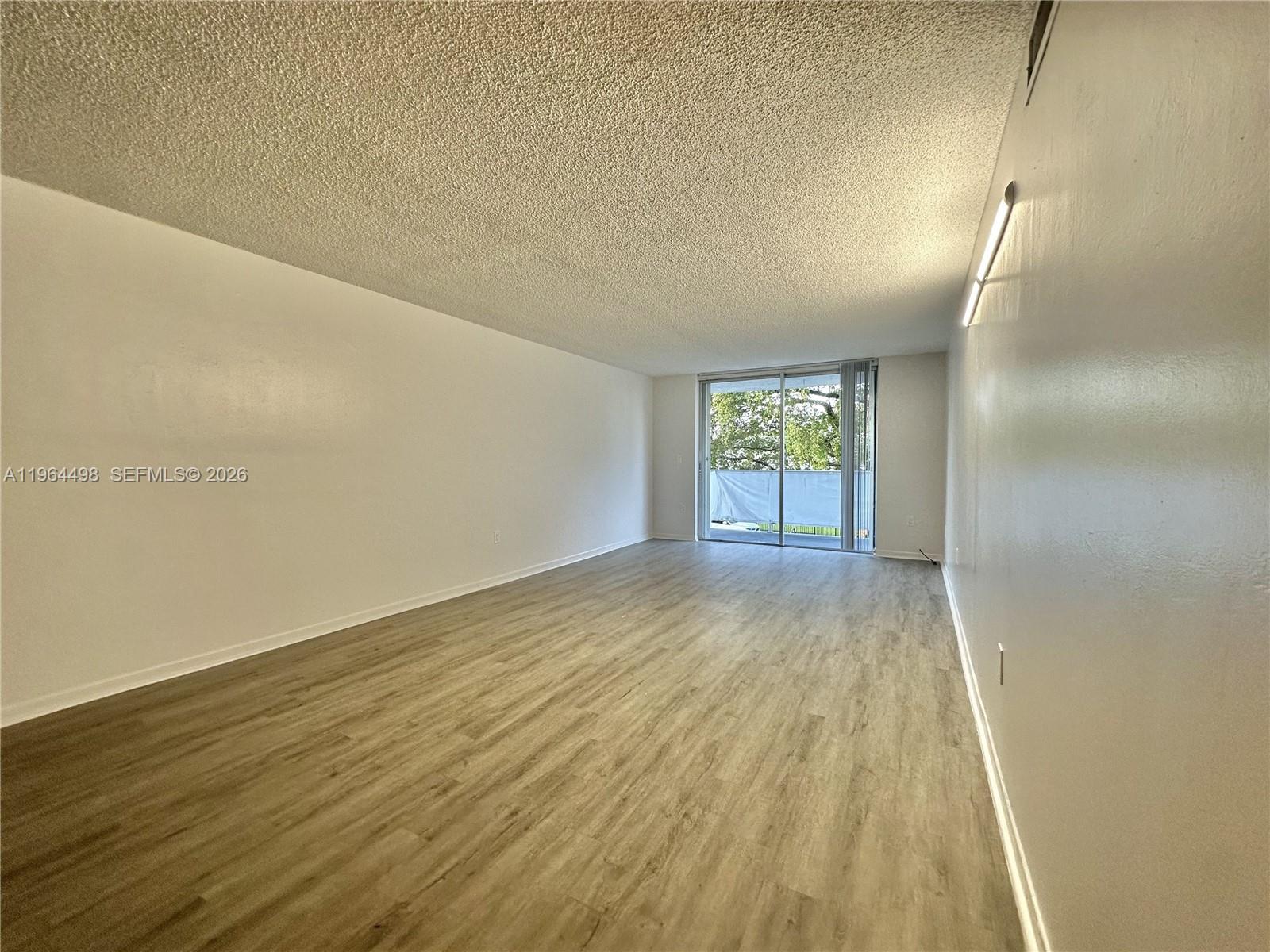 An empty living room features light wood-look flooring, white walls, and a sliding glass door — 1470 NE 125th Ter # 210, North Miami FL 33161, NORTH MIAMI, FL