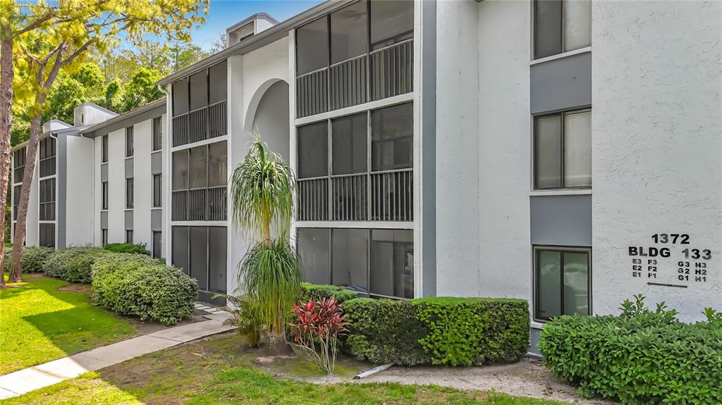 A three-story apartment building exterior features screened balconies, white stucco walls, and landscaped bushes — 1372 PINE RIDGE CIRCLE E UNIT H3, TARPON SPRINGS, FL