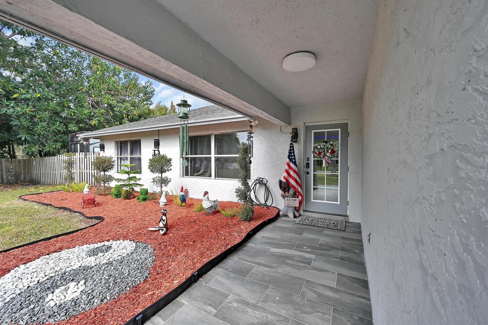 A covered porch with gray tile flooring leads to a front door with a flag — 2404 Dade Road, Fort Pierce FL 34982, FORT PIERCE, FL