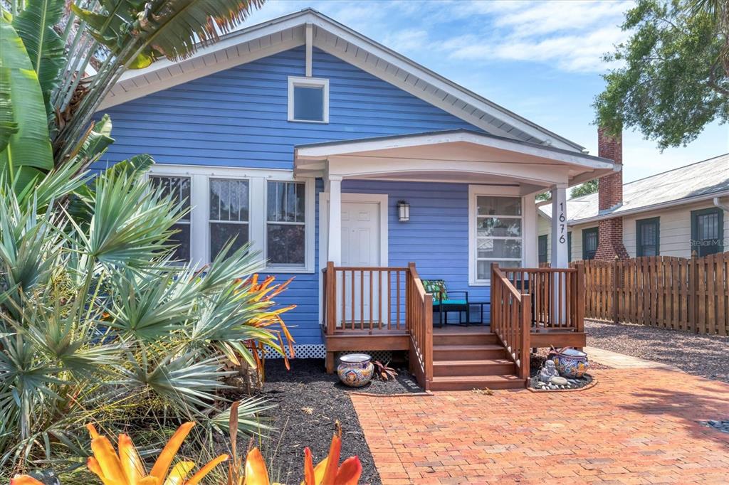 A blue house with a wooden porch and brick walkway stands behind a tropical garden — 1676 OAK STREET, SARASOTA, FL