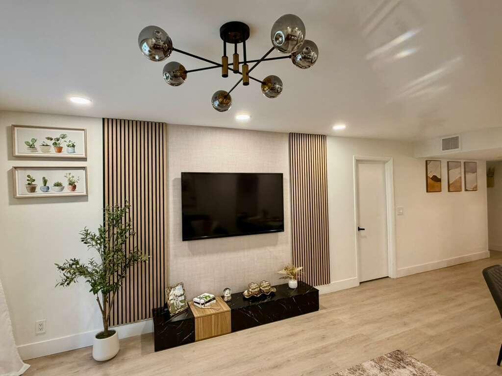 A living room features a wall-mounted television, vertical wood slat panels, and a modern chandelier — 22415 SW 61st Way # 106, Boca Raton FL 33428, BOCA RATON, FL