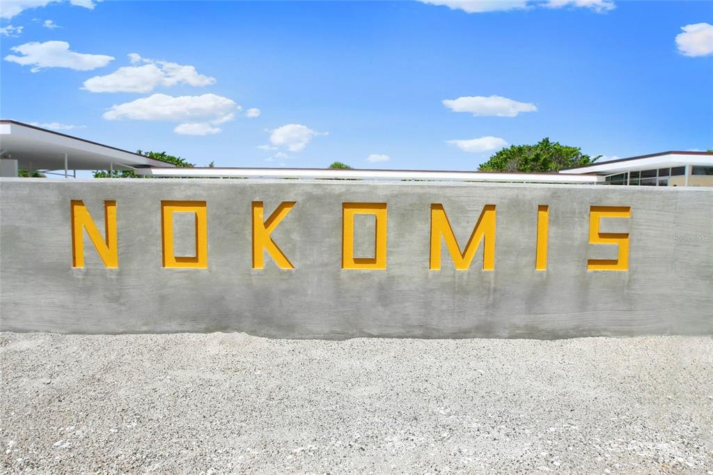 A concrete wall displays the yellow-lettered name NOKOMIS under a clear blue sky — 116 AVENIDA LA PALMA, NOKOMIS, FL