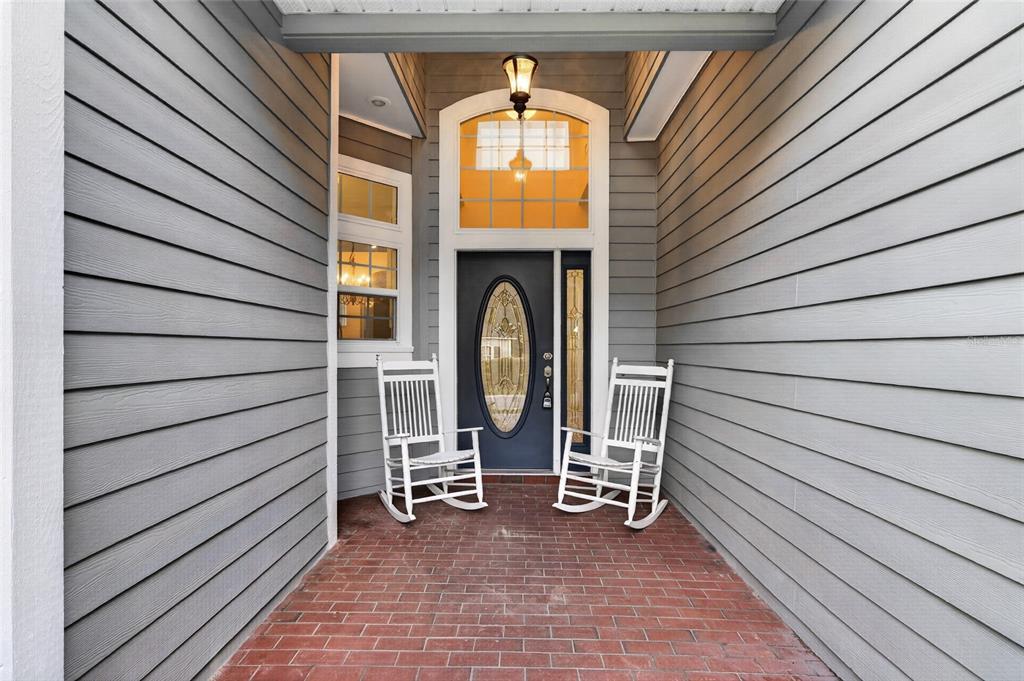 A covered brick porch features two white rocking chairs in front of a dark front door — 914 SW 83RD TERRACE, GAINESVILLE, FL