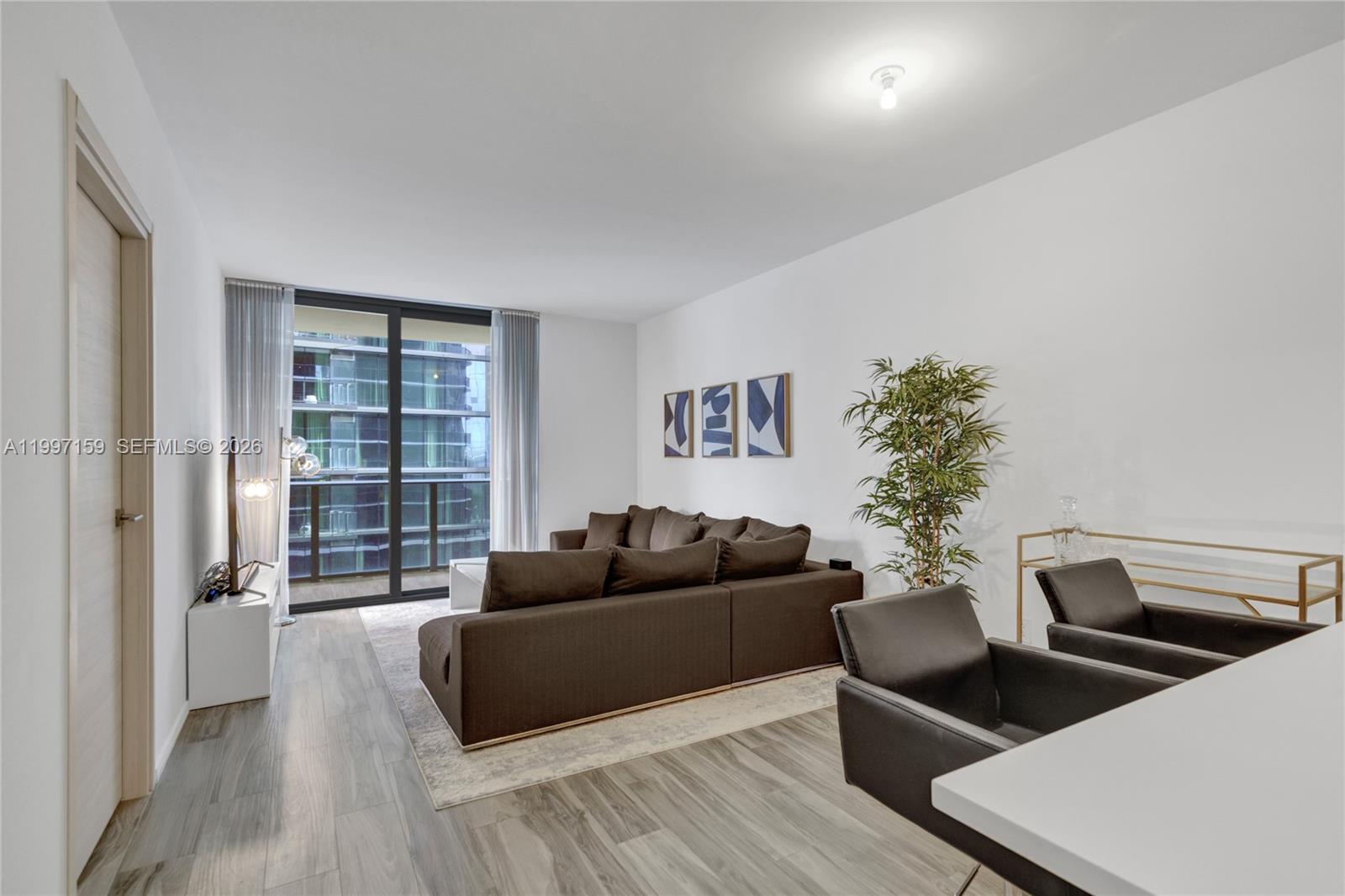 A living room features a brown sectional sofa, wood-look flooring, and a balcony with city views — 801 S Miami Ave # 1106, Miami FL 33130, MIAMI, FL