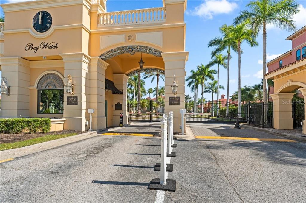 The entrance to a gated community features a beige archway with a clock and palm trees — 2747 VIA CAPRI UNIT 1132, CLEARWATER, FL