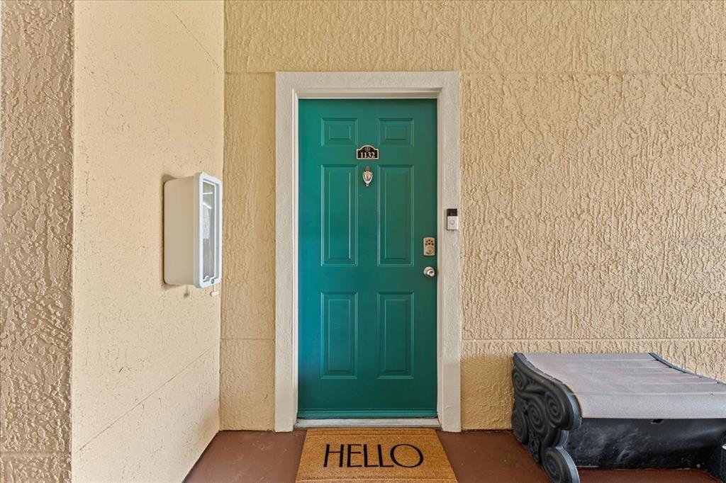 A teal front door with a welcome mat sits on a beige stucco porch — 2747 VIA CAPRI UNIT 1132, CLEARWATER, FL