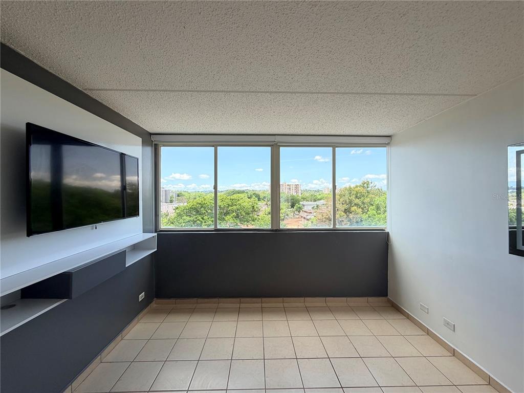 A room with light tile floors, a wall-mounted television, and large windows overlooking trees — #850 CALLE EIDER UNIT A808, SAN JUAN, PR