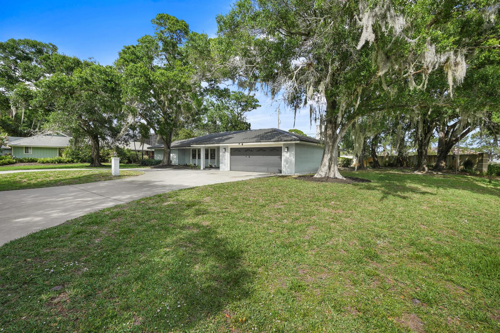 A light-colored single-story house with a gray garage door sits behind a large grassy lawn — 7312 Elyse Circle, Port St. Lucie FL 34952, PORT ST. LUCIE, FL