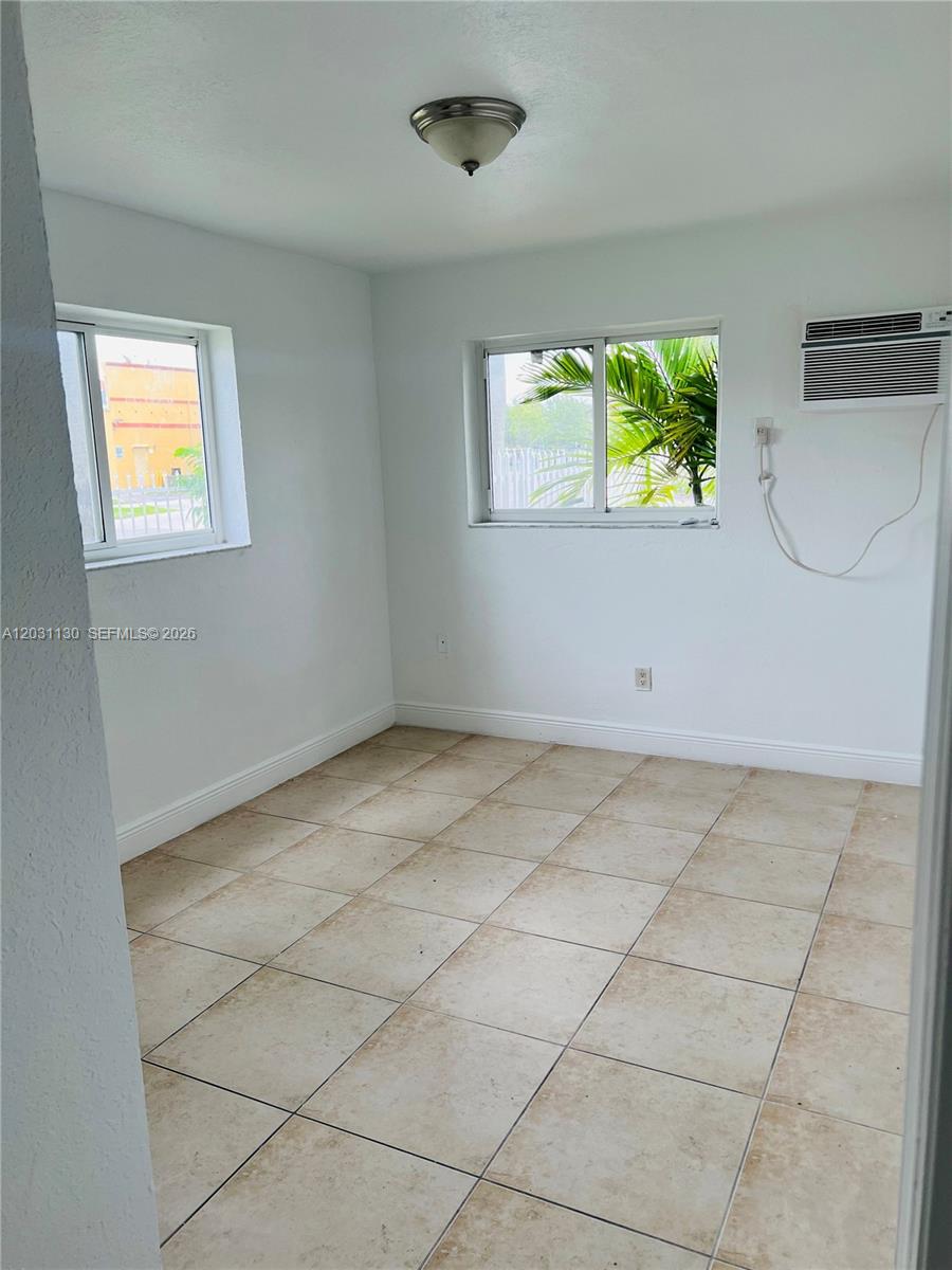 An empty room features white walls, light-colored tile flooring, two windows, and a wall-mounted air conditioner — 880 NE 18th Ave # 3, Homestead FL 33033, HOMESTEAD, FL