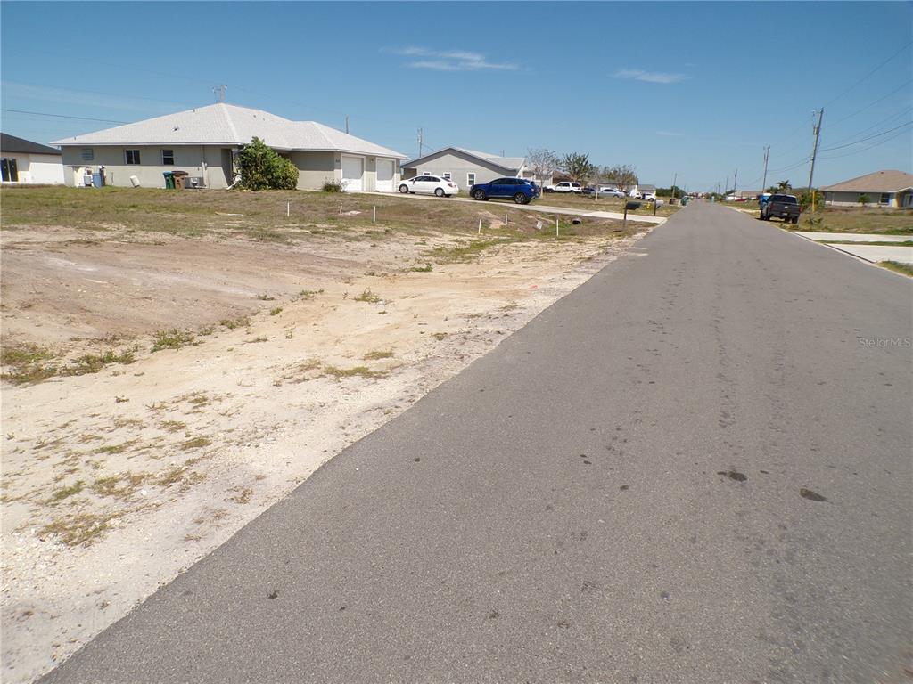A paved road runs alongside a vacant sandy lot with houses in the background — 825 NE 15TH LANE, CAPE CORAL, FL