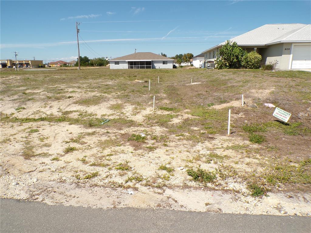 A vacant, grassy residential lot features several white survey stakes and a plumbing company sign — 825 NE 15TH LANE, CAPE CORAL, FL