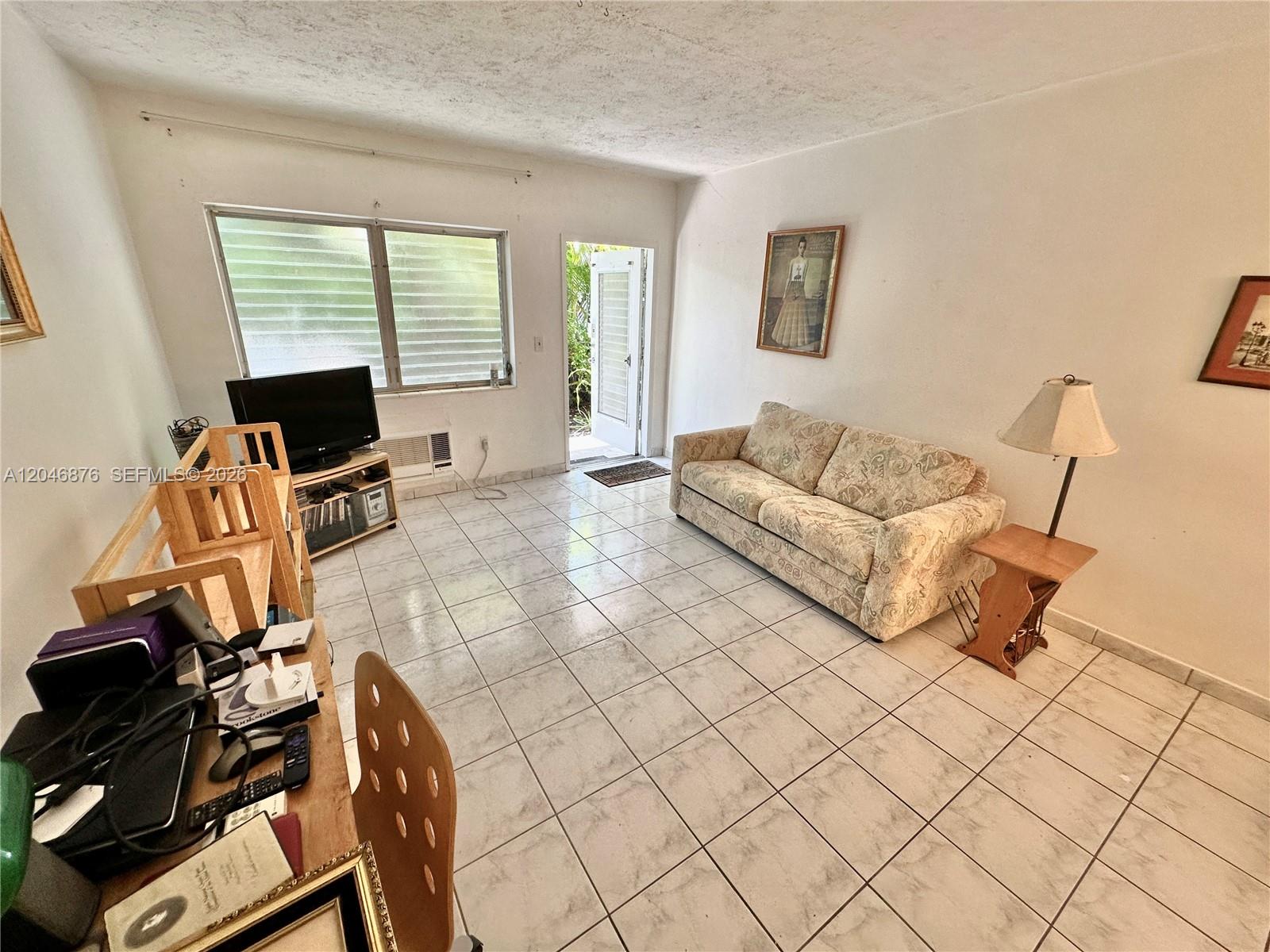 A living room features white walls, tiled floors, a patterned sofa, and an exterior door — 8132 Harding Ave # 6, Miami Beach FL 33141, MIAMI BEACH, FL