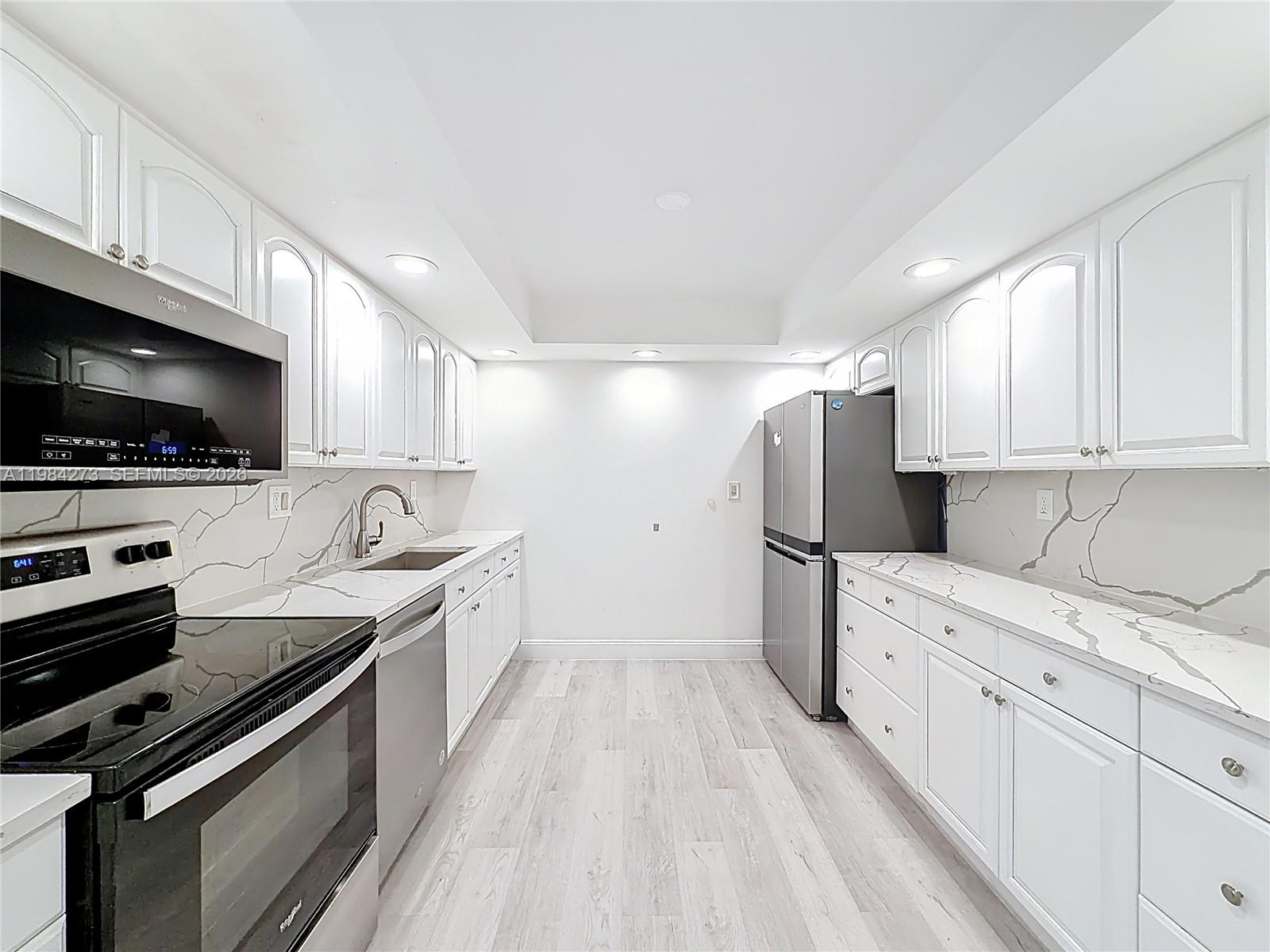 A galley kitchen features white cabinets, marble-patterned countertops, stainless steel appliances, and light wood flooring — 1000 Parkview Dr # 121, Hallandale Beach FL 33009, HALLANDALE BEACH, FL