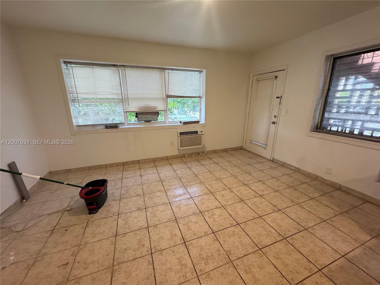 An empty room features beige tile flooring, a large window with blinds, and a wall-mounted AC — 7300 Harding Ave # 21, Miami Beach FL 33141, MIAMI BEACH, FL