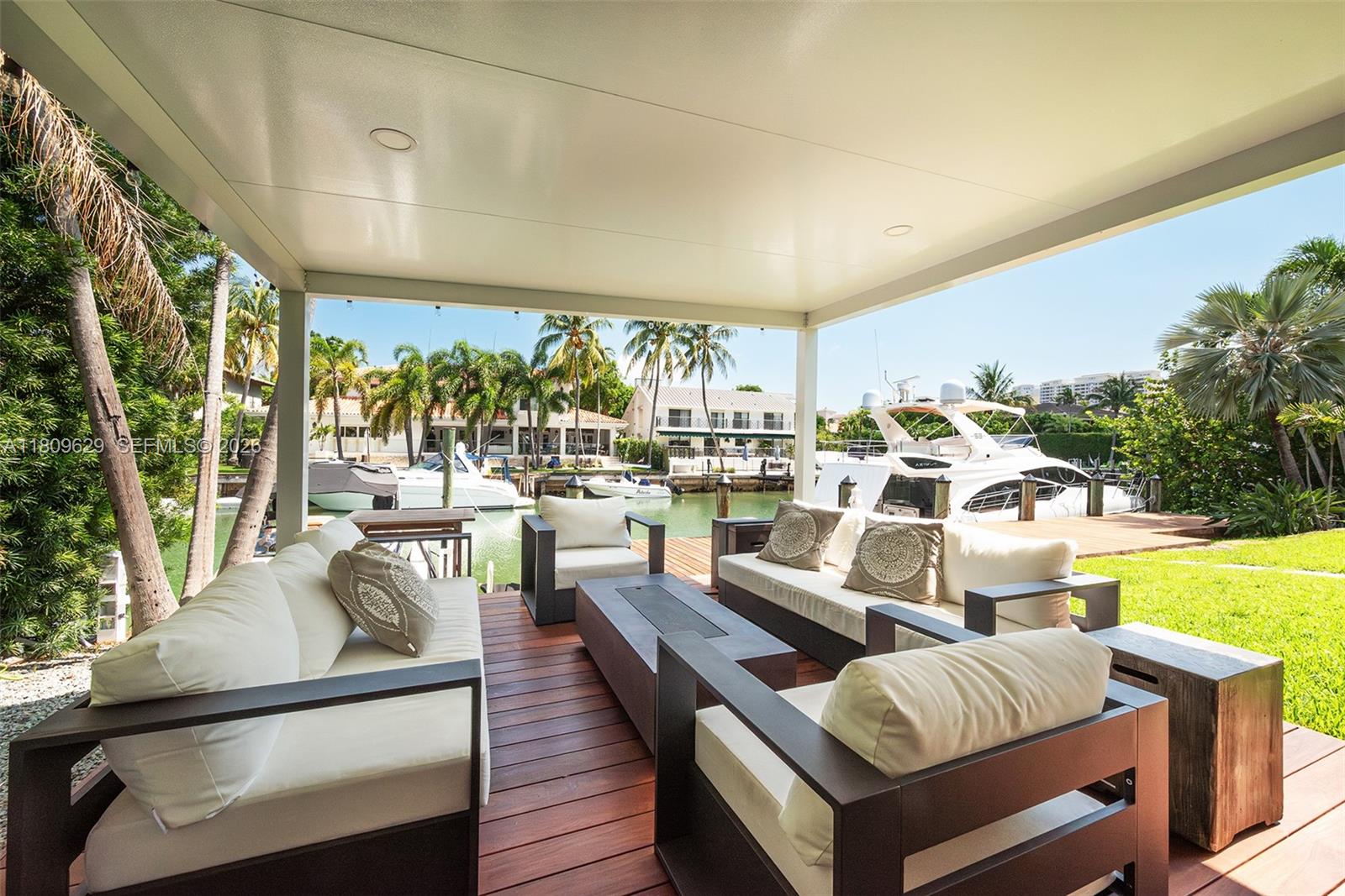 A covered outdoor patio features modern furniture on a wooden deck overlooking a boat canal — 201 Knollwood Dr, Key Biscayne FL 33149, KEY BISCAYNE, FL