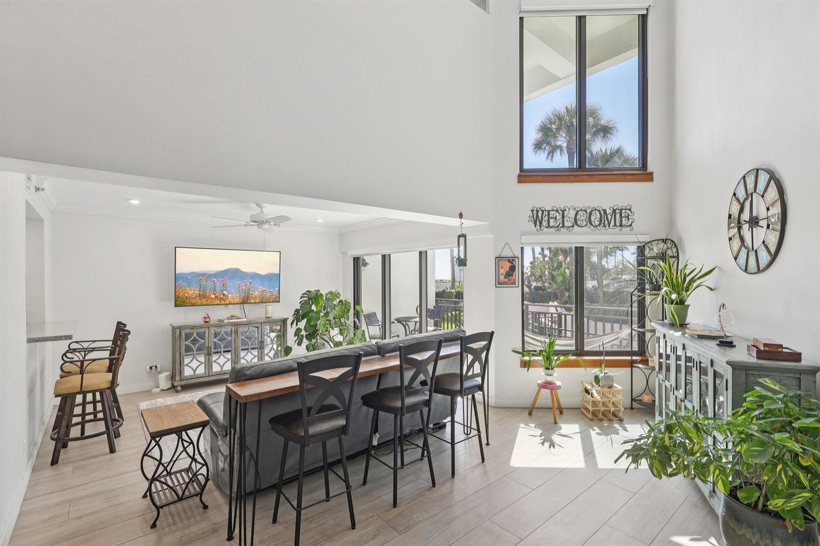 A bright living room features a sofa with a bar table, television, and large windows — 665 SE 21st Avenue # 107, Deerfield Beach FL 33441, DEERFIELD BEACH, FL