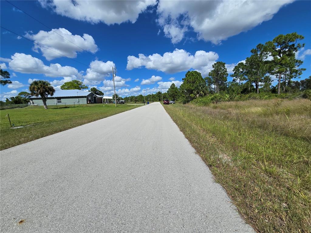 A paved road stretches between a grassy lot and a house under a cloudy sky — 1034 DEARBORN STREET, LEHIGH ACRES, FL
