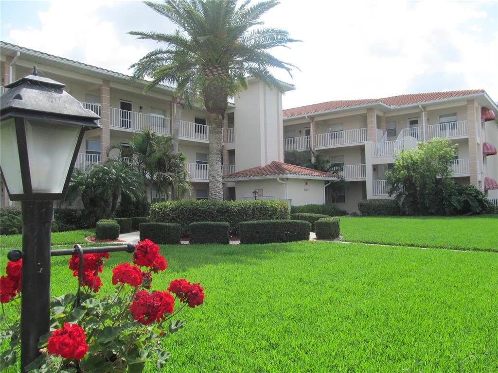 A multi-story apartment building with a palm tree, manicured hedges, and red flowers in front — 6507 STONE RIVER ROAD UNIT 210, BRADENTON, FL