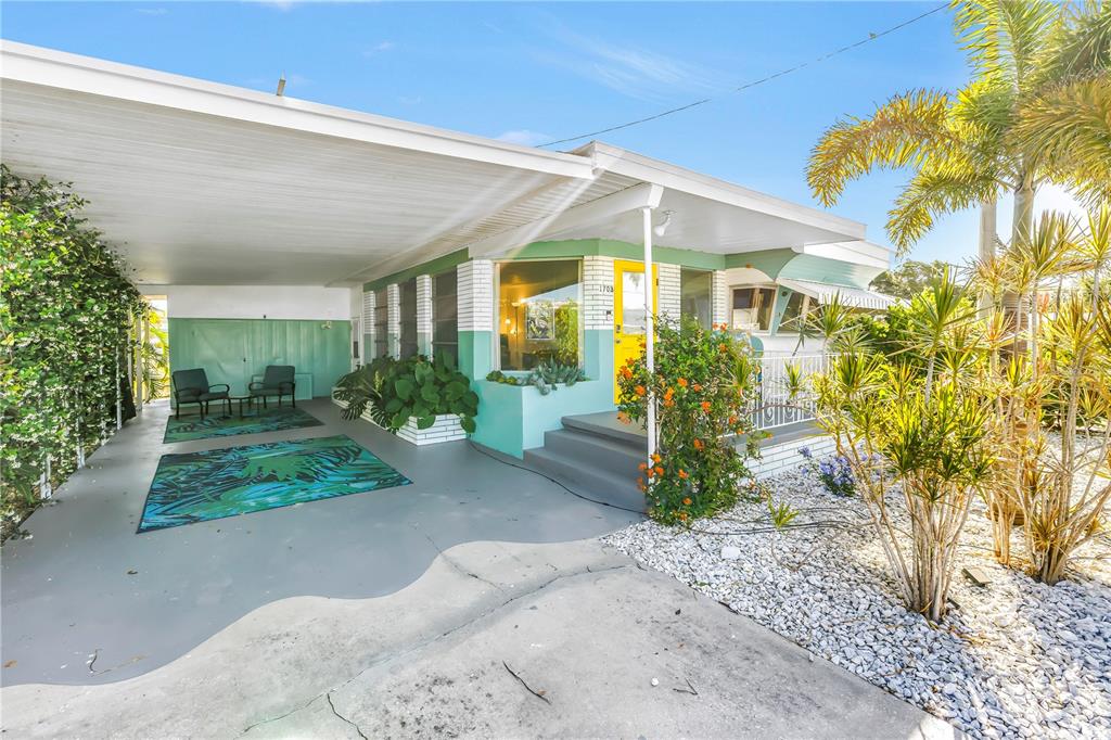 A single-story house features a carport, a bright yellow front door, and tropical landscaping — 1703 MINNESOTA AVENUE, BRADENTON, FL