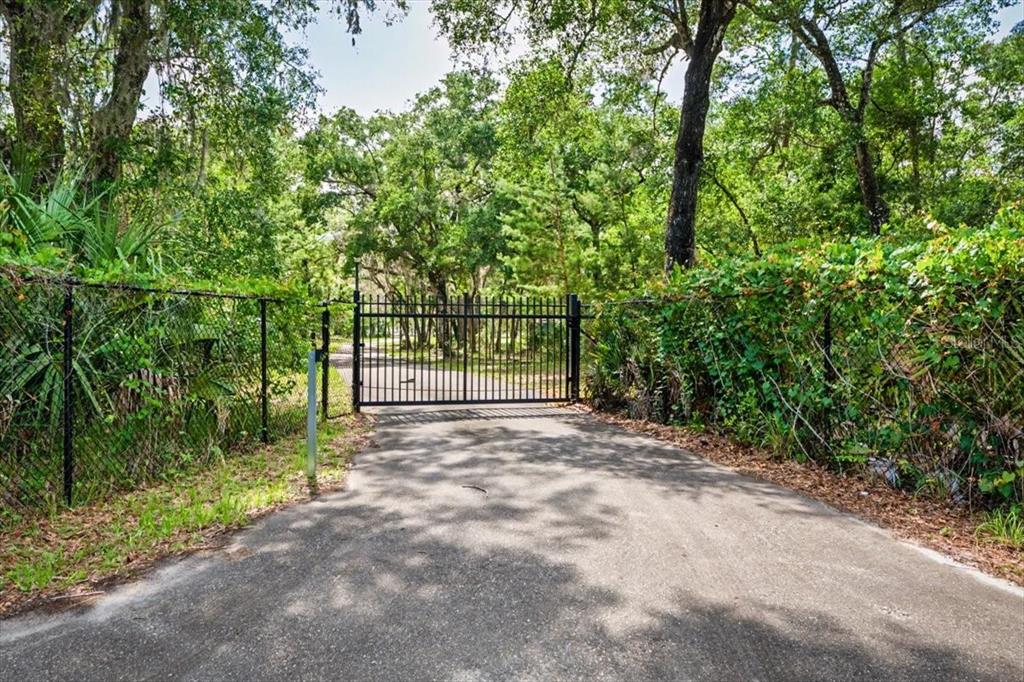 A black metal gate blocks a paved driveway surrounded by trees and chain-link fencing — 1812 TAYLOR ROAD, PORT ORANGE, FL