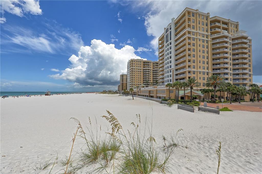 A wide sandy beach stretches toward tall residential buildings under a partly cloudy blue sky — 10 PAPAYA STREET UNIT 305, CLEARWATER BEACH, FL