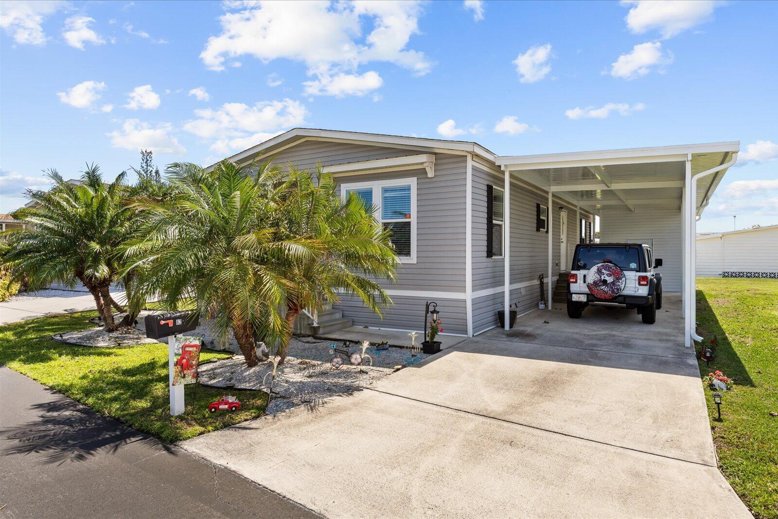 A gray manufactured home features a concrete driveway, a carport, and a palm tree — 82 Sandhill Boulevard, Fort Pierce FL 34982, FORT PIERCE, FL
