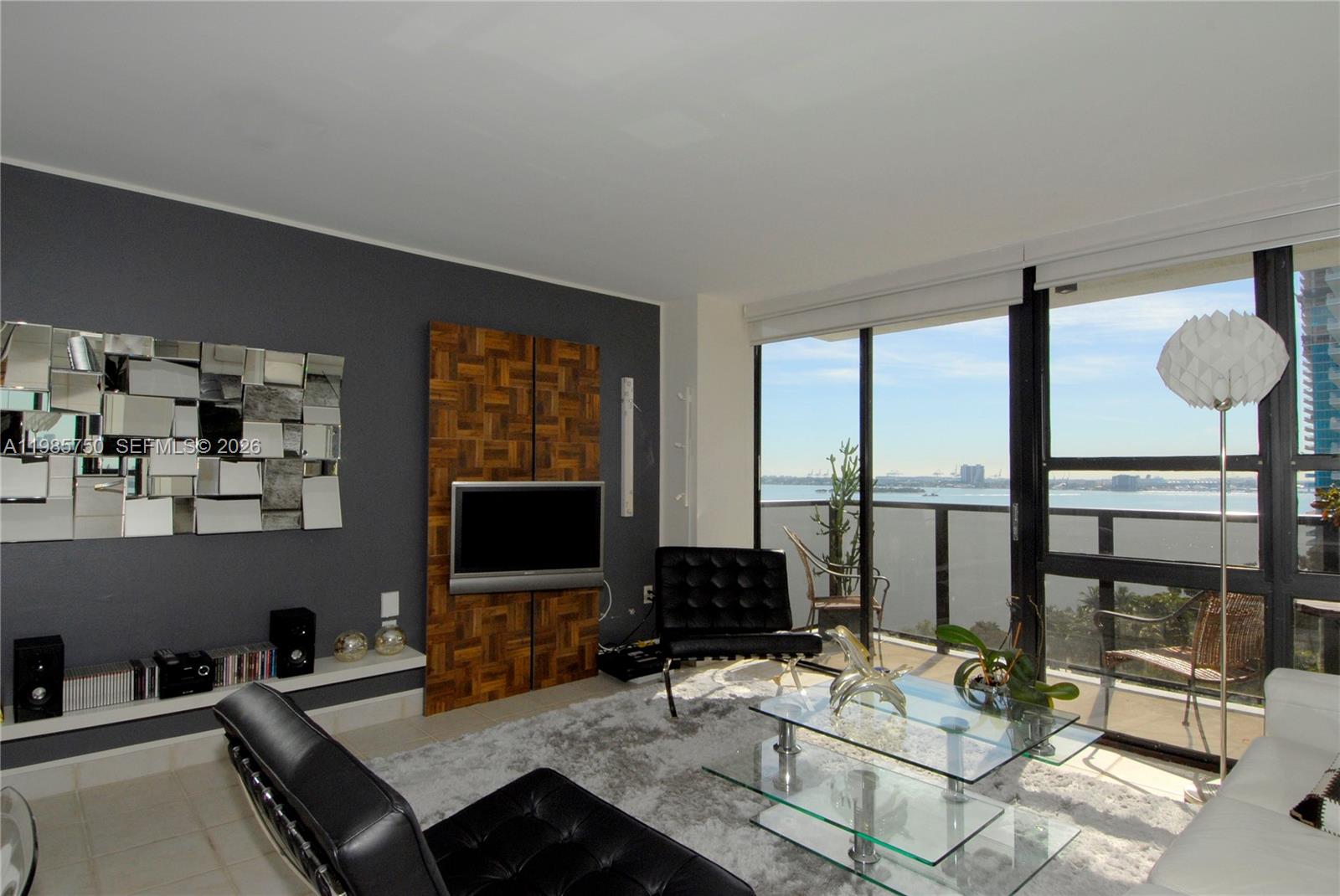 A modern living room features dark gray walls, a glass coffee table, and balcony water views — 600 NE 36th St # 1212, Miami FL 33137, MIAMI, FL
