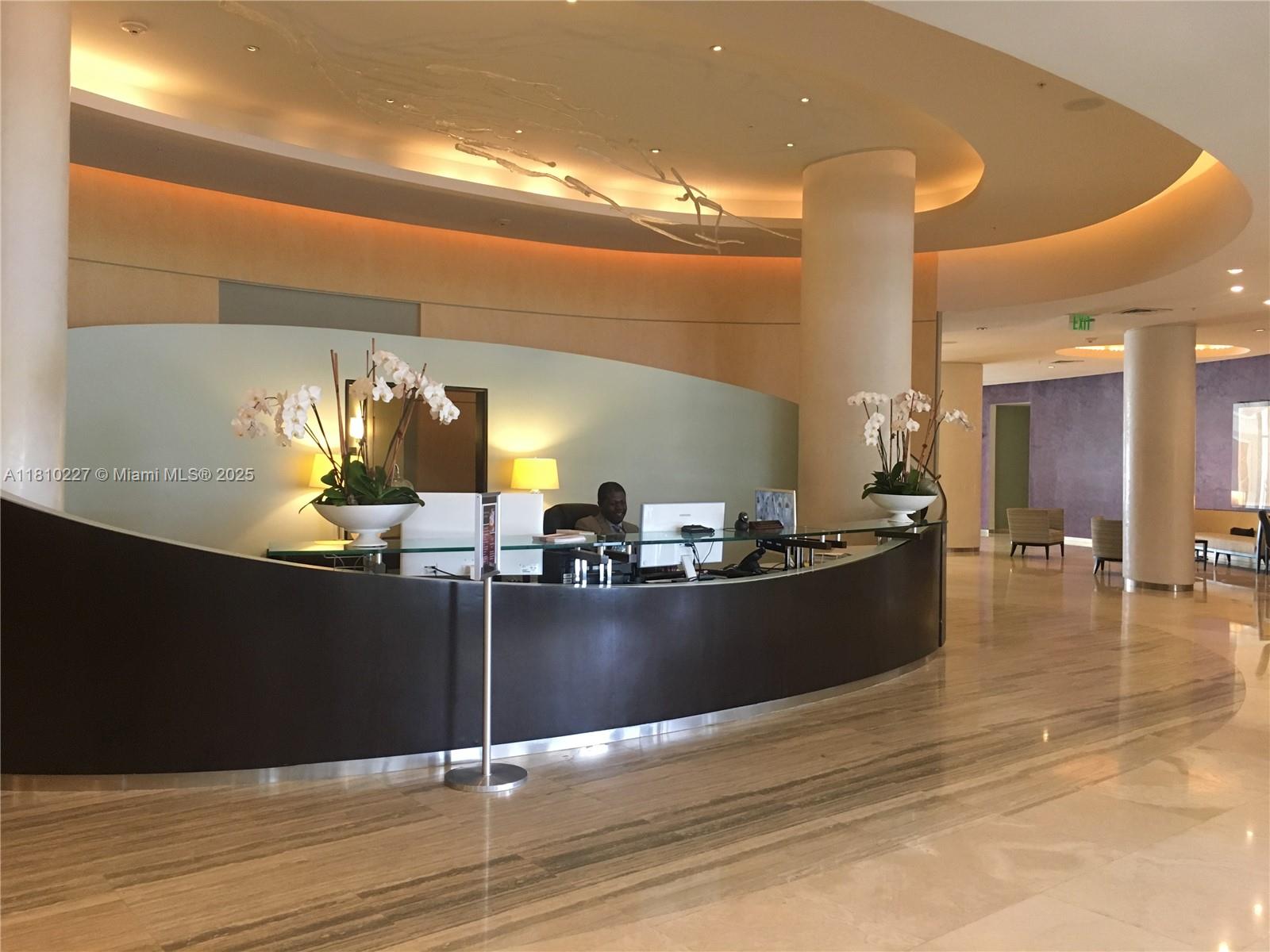 A modern lobby features a curved reception desk, marble flooring, and decorative ceiling lighting — 10275 Collins Ave # 628, Bal Harbour FL 33154, BAL HARBOUR, FL