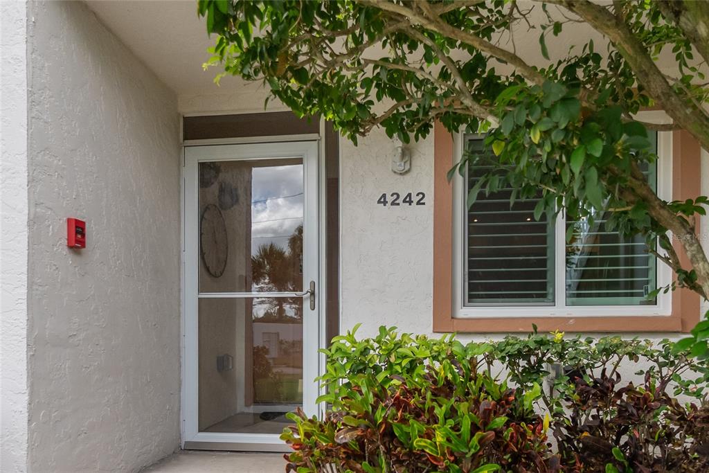 A white front door with a storm door is next to a window and landscaping — 4242 MADEIRA COURT UNIT 3315, SARASOTA, FL