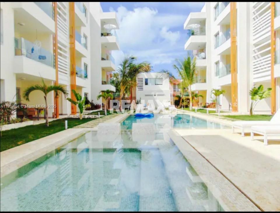 A swimming pool sits between two modern white apartment buildings with balconies and palm trees — El cortesito Punta Cana # 1, PU, PU