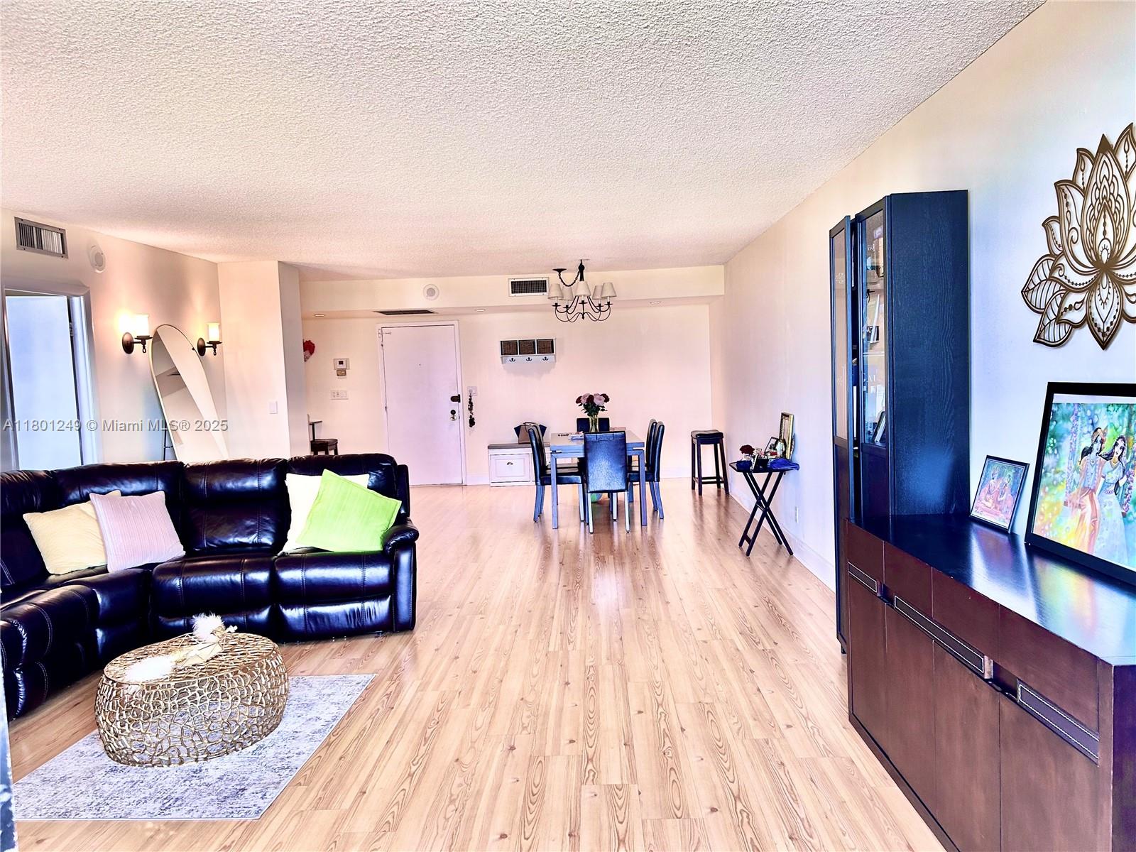 This open-concept living and dining area features wood-look flooring and a dark leather sectional sofa — 600 Three Islands Blvd # 905, Hallandale Beach FL 33009, HALLANDALE BEACH, FL