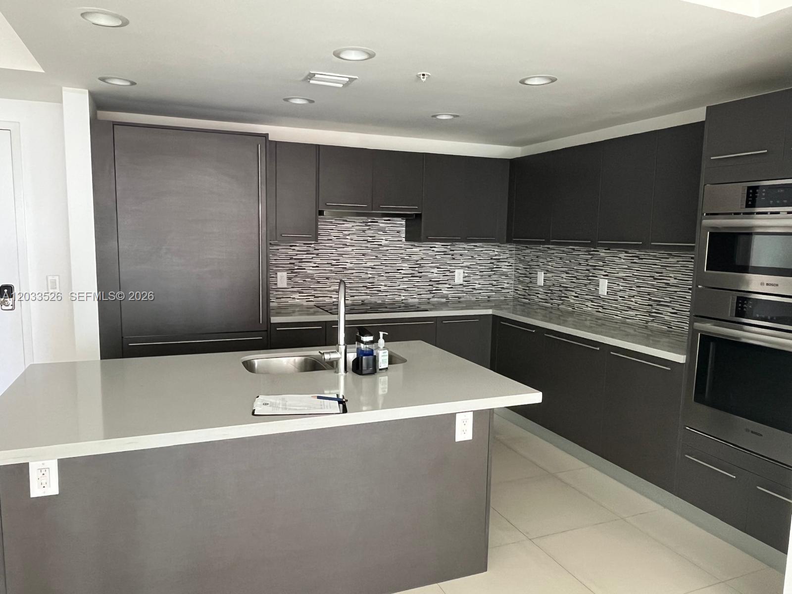 A modern kitchen features dark gray cabinetry, a central island, and a horizontal-patterned tile backsplash — 7825 NW 107th Ave # 506, Doral FL 33178, DORAL, FL