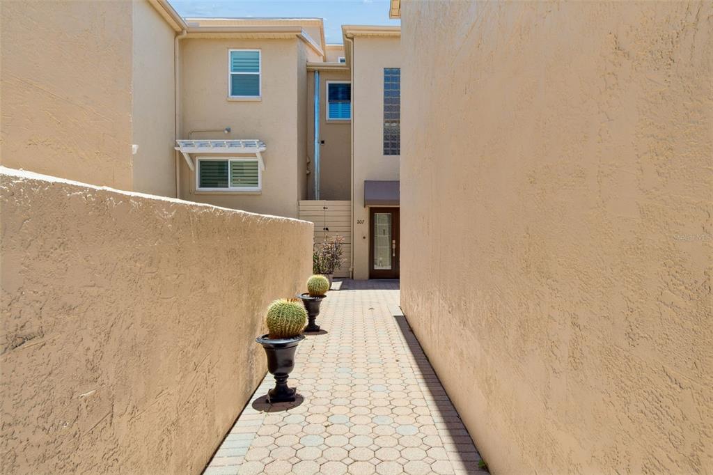A narrow paved walkway leads between two stucco walls toward a residential building entrance — 487 PINELLAS BAYWAY S UNIT 207, ST PETERSBURG, FL