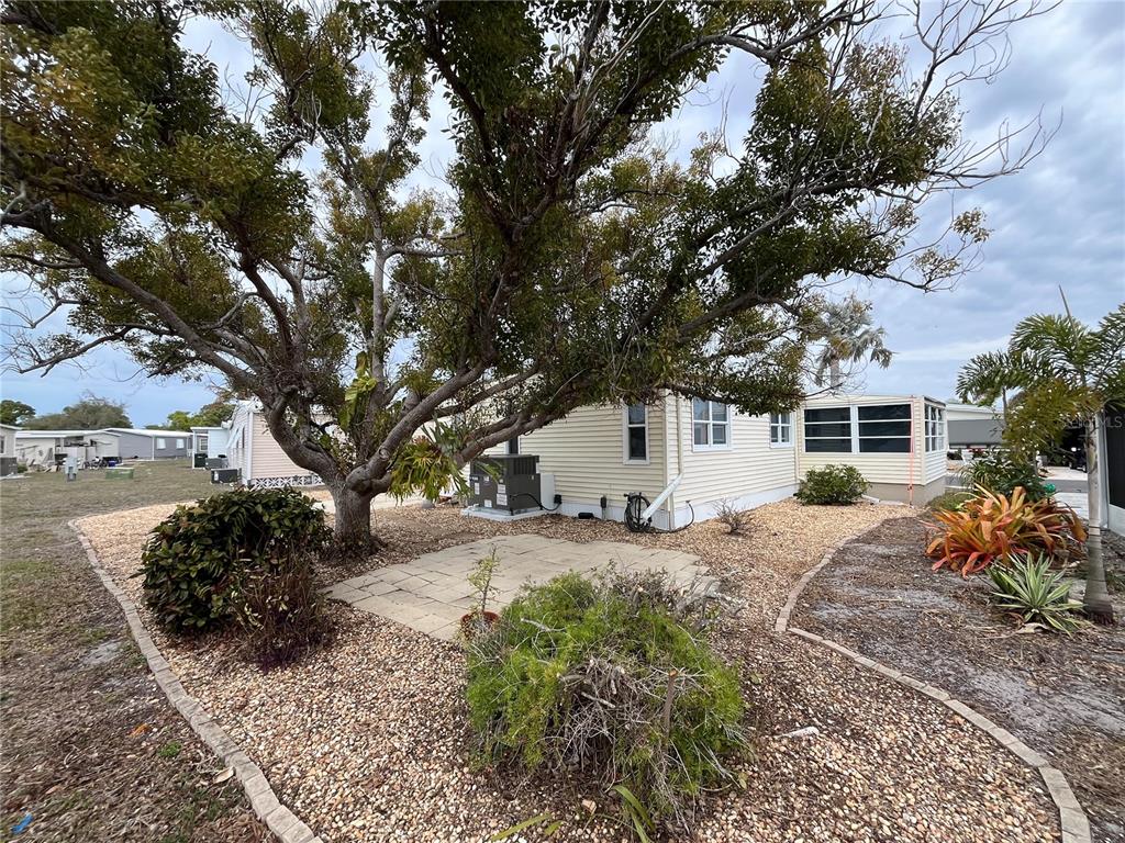 A large tree shades a small stone patio next to a light-colored mobile home — 873 BAVENO DRIVE, VENICE, FL